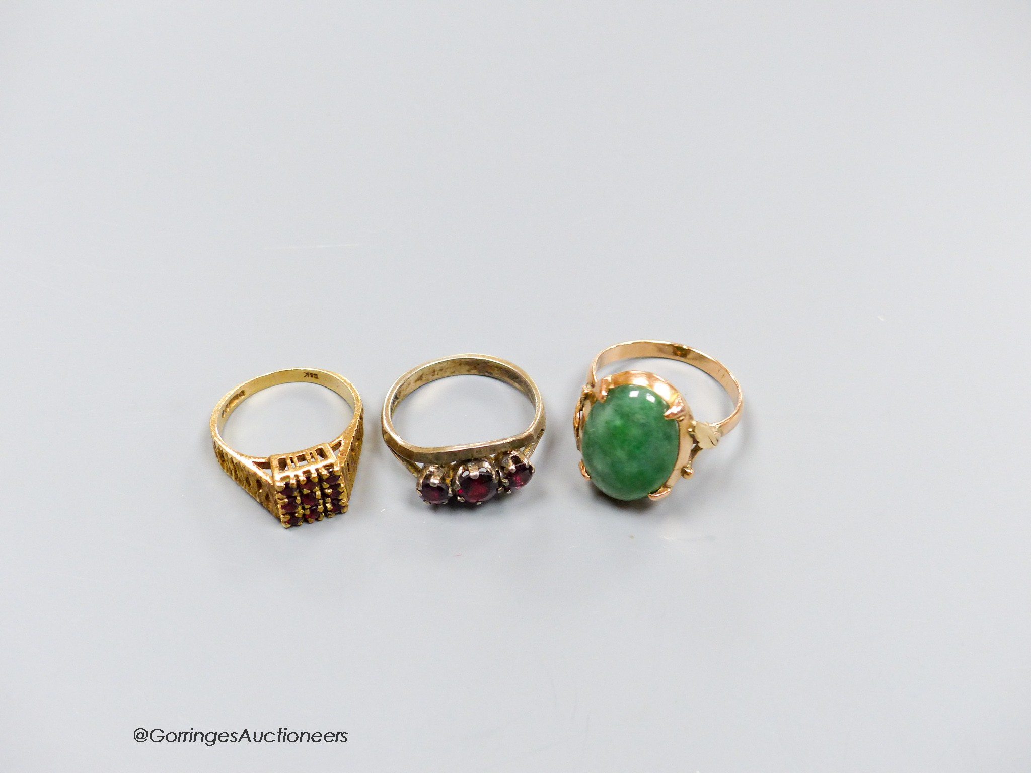 A 9ct gold and garnet cluster ring, a similar white metal ring and a yellow metal and jadeite cabochon ring, gross weight 12.1 grams.